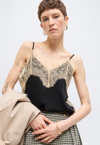 Νεαρό άτομο με κοντά μαλλιά φοράει μαύρο καμιζολάκι με δαντέλα, φούστα με σχέδια, χρυσά σκουλαρίκια, βραχιόλι και κρατάει μπεζ σακάκι σε λευκό φόντο.