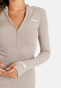 Body de manga larga ribeteado en color beige, con un cierre de media cremallera en la parte frontal y el nombre de la marca bordado en la manga; diseño ajustado, textura suave.
