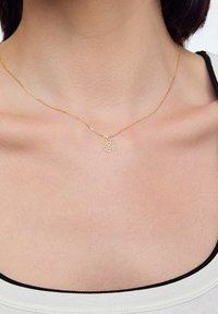 Złoty naszyjnik z delikatnym łańcuszkiem i okrągłym wisiorkiem z wzorem wyciętym w kształcie kwiatu. Powierzchnia jest gładka i błyszcząca.