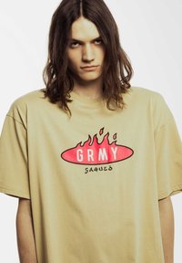 Camiseta oversize beige con un gráfico ovalado rojo y letras blancas "GRMY" con acentos de llamas, hecha de material de algodón suave.