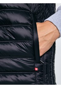 Fényes szürke ujjú kéz fekete steppelt dzseki zsebében, kis piros címkével, amelyen fehér csillag látható.