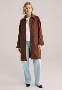 Palton maro din piele întoarsă cu guler, nasturi în față și croială lejeră, asortat cu blugi albastru deschis și pantofi negri cu vârf ascuțit. Cămașă albă simplă pe dedesubt.