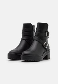 Botas negras de tobillo hechas de faux cuero suave, con correas ajustables, un tacón grueso y una suela de goma texturizada.