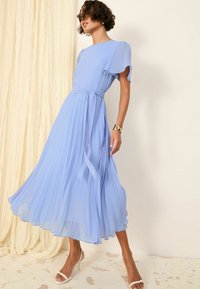 Robe plissée bleu clair avec des manches courtes et flottantes, taille cintrée et jupe fluide. Se termine au-dessus des chevilles, associée à des talons blancs.