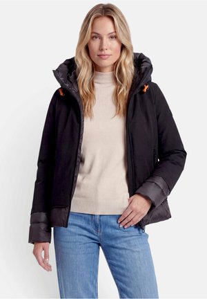 Femme aux longs cheveux blonds portant une veste noire à capuche par-dessus un pull beige et un jean bleu, debout devant un fond clair uni.