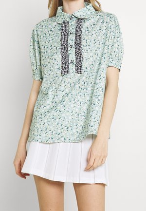 Femme portant un chemisier fleuri vert menthe avec patte de boutonnage à volants vichy noir et une jupe plissée blanche, debout sur fond blanc.