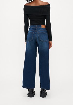 Femme portant un jean bleu foncé à jambes larges, un haut noir à manches longues et épaules dénudées, et des bottes noires épaisses, debout de dos sur fond blanc.