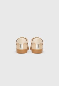 Baskets avec une tige en cuir beige, accents métalliques dorés sur le talon et une semelle en caoutchouc texturée, présentant un design à profil bas.