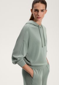 Mentazöld kapucnis pulóver bordás részletekkel és laza szabással. Az anyag sima megjelenésű, puha tapintású. Elülső zsebek láthatók.