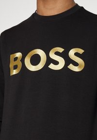 Svart långärmad skjorta med djärva, metalliska guldbokstäver som stavar "BOSS" över bröstet, som visar en stilren och modern design.