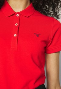 Femme portant un polo rouge à manches courtes avec trois boutons et un petit logo GANT sur la poitrine gauche.