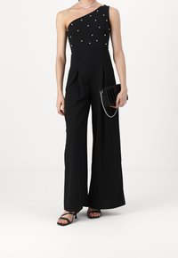 Zwarte jumpsuit met één schouder, voorzien van parelversieringen, brede pijpen en een textuurstof. Gecombineerd met een geplooide zwarte clutch.