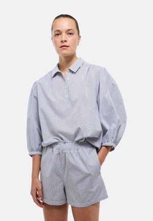 Femme à la peau claire et aux cheveux attachés porte un chemisier ample à carreaux bleu clair avec des manches trois-quarts et un short assorti à taille élastique.