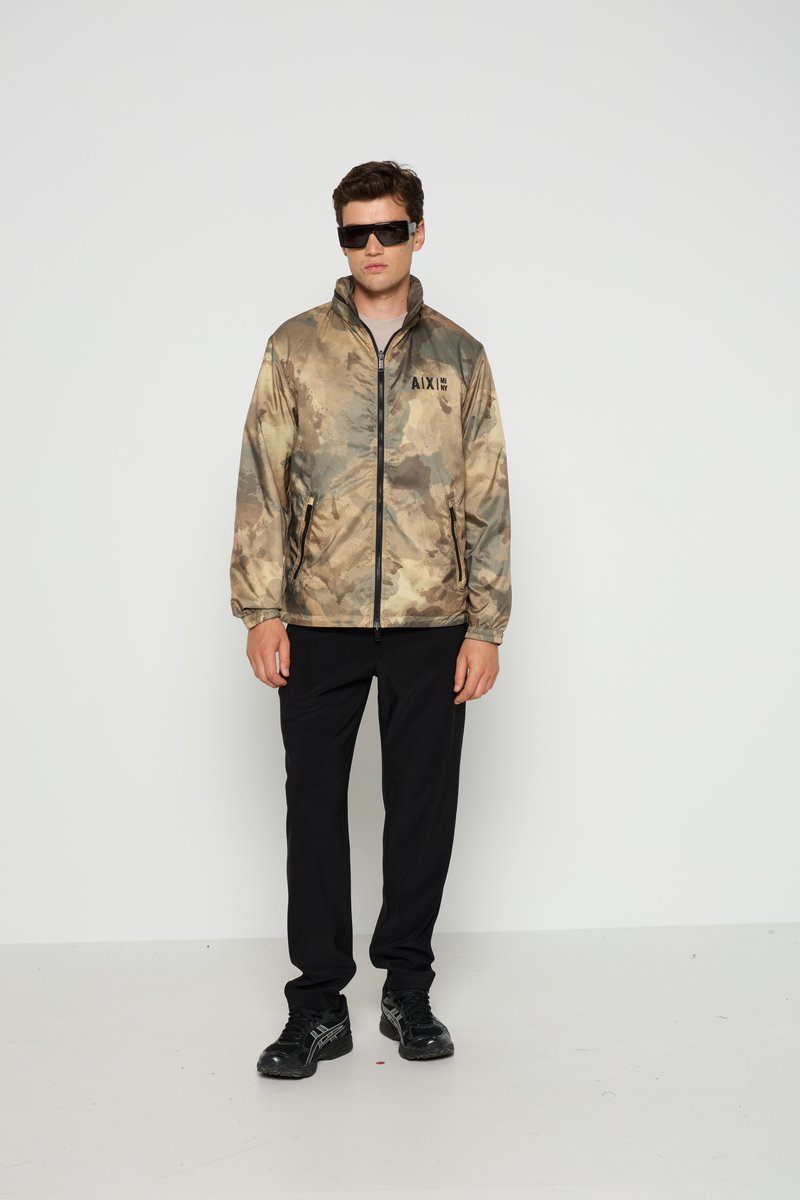 Camouflagejack in beige en olijfgroene tinten, met een opstaande kraag, ritsluiting en branding op de borst. Gepaard met zwarte broek en schoenen.