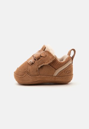 Chaussure bébé en daim marron avec doublure en peluche, languette à boucle au talon et détail noeud sur le côté sur fond clair uni.