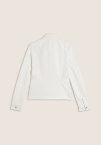 Veste ajustée blanche, tissu doux, manches longues, col à revers, coutures au dos pour la forme, et accents de boutons sur les détails des poignets.