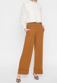 Blouse courte en dentelle blanche avec des manches longues et un col haut, associée à un pantalon large marron avec des revers et des poches latérales.