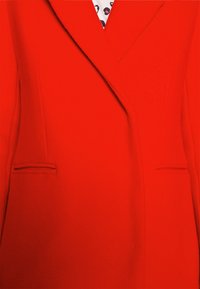 Manteau rouge vif sur mesure avec revers et poches passepoilées, laissant partiellement apparaître une chemise blanche à imprimé léopard noir et rose en dessous.