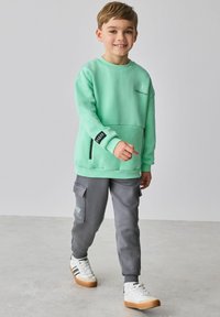 Mintgroene trui met kangoeroezak, grijze joggingbroek met steekzakken en witte sneakers met bruine zolen. Kind loopt op een lichte ondergrond.
