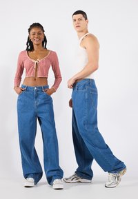 Red checkered cropped top with a bow detail, paired with blue wide-leg denim jeans. Model wears multicolored sneakers, standing against a white backdrop.