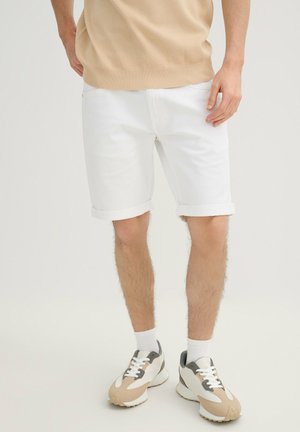 Hombre con camiseta de punto beige, pantalones cortos blancos remangados, calcetines blancos y zapatillas beige blanco gris, de pie sobre fondo claro.