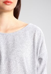 Ljusgrå, mjuk stickad tröja med lös passform och vid halsringning. Slät textur, minimal mönstring och långa ärmar.