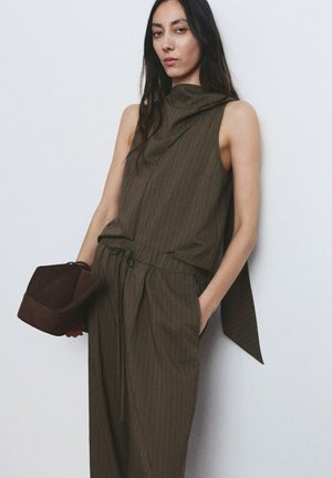 Frau mit langem, dunklem Haar, die einen ärmellosen dunkelolivgrünen, dünn gestreiften Overall trägt, eine dunkelbraune Handtasche hält und vor einer weißen Wand steht.