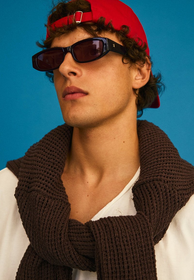 Jeune personne aux cheveux bouclés porte des lunettes de soleil noires, une casquette rouge portée à l'envers, une chemise blanche et une écharpe en tricot marron foncé sur fond bleu.
