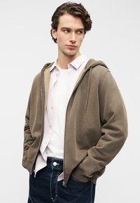 Καφέ φούτερ με φερμουάρ, με μαλακή εσωτερική επένδυση, μπροστινές τσέπες και κορδόνια. Φοριέται πάνω από ένα λευκό πουκάμισο με ανοιχτό ροζ πουκάμισο με κουμπιά από κάτω.