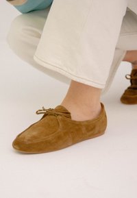 Scarpe slip-on in suede marrone con punta arrotondata, caratterizzate da un dettaglio a fiocco anteriore e suola piatta, abbinate a pantaloni croppati di colore chiaro.
