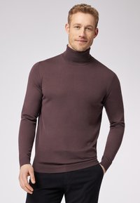Rollkragenpullover in dunklem Mauve, gerippte Textur, schlanker Schnitt, lange Ärmel und ein enganliegender Kragen. Getragen über dunklen Hosen.