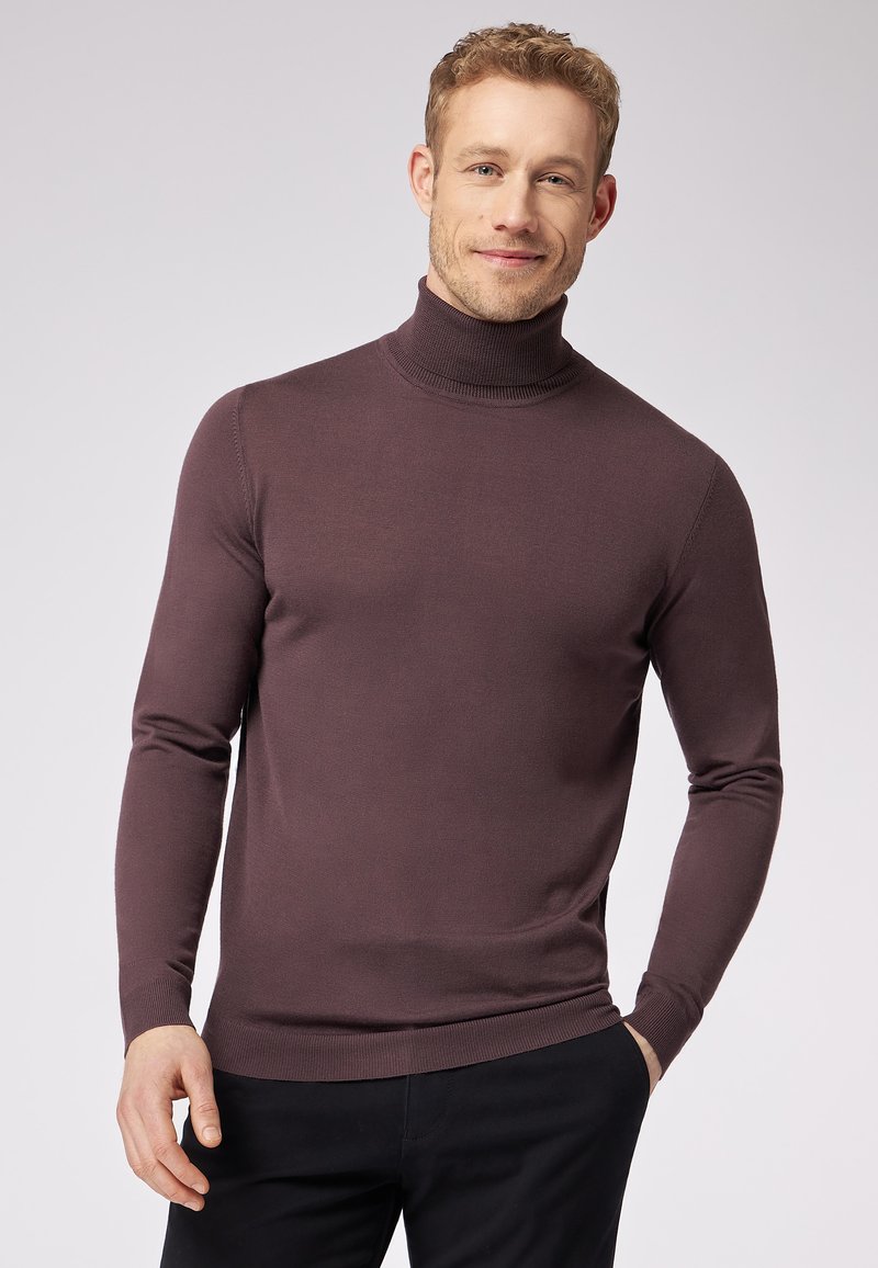Rollkragenpullover in dunklem Mauve, gerippte Textur, schlanker Schnitt, lange Ärmel und ein enganliegender Kragen. Getragen über dunklen Hosen.
