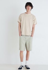 Beige pamut póló, világoszöld rövidnadrág és fekete/fehér lábbeli. A modell áll, kezét a zsebébe dugva egy sima fehér háttér előtt.