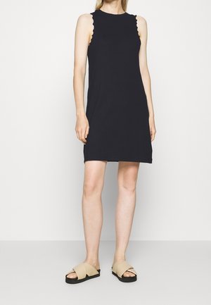 Kobieta ubrana w czarną, bezrękawną sukienkę z falistymi krawędziami oraz beżowe sandały wsuwane, stojąca na białym tle.