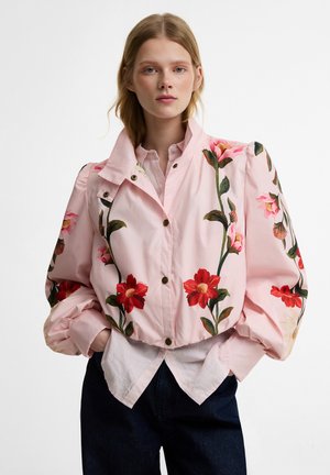 Vrouw die een roze jas draagt met grote bloemrijke borduursels, pofmouwen en handen in de zakken, gedragen over een roze hemd en donkere broek.