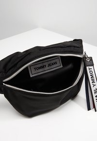 Svart tygfannypack med en silverdragkedja, med en "TOMMY JEANS" etikett inuti. Inkluderar ett band med röda, vita och blå accenter.