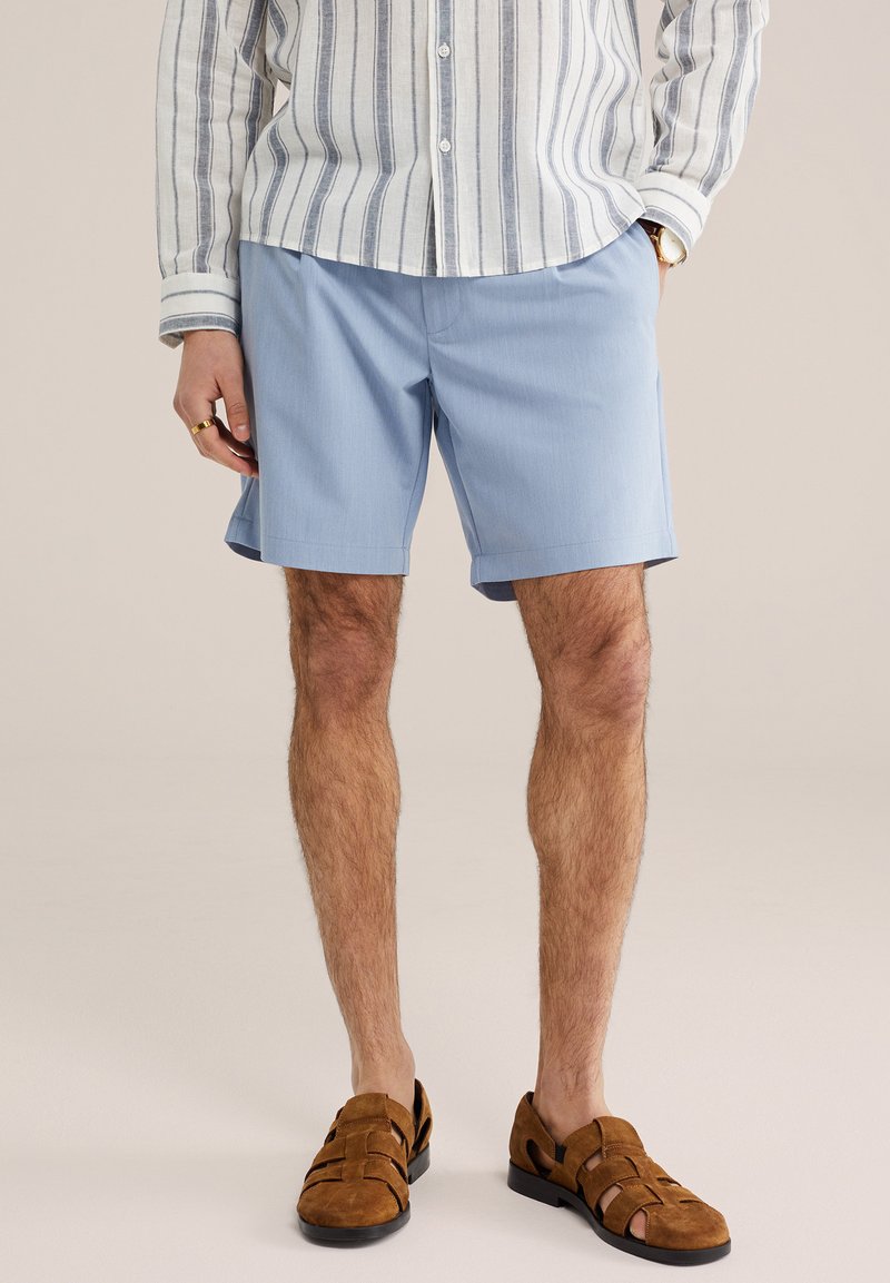 Homme portant un short bleu clair, une chemise à manches longues rayée, des sandales habillées en daim marron, une montre en or et une bague en or.