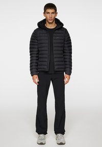 Kvadderad svart jacka med huva, som har dragkedjestängning och horisontell quiltad design. Buren med en svart skjorta och svarta byxor. Grå sneakers.