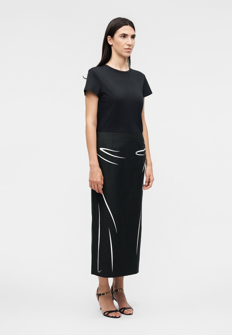 Woman stands facing forward wearing a black short-sleeve shirt and a black midi skirt with white abstract line designs and black heels.