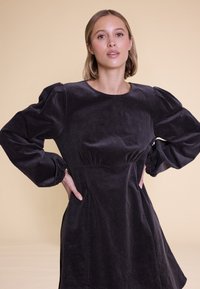 Puff-sleeved, mørkegrå kordfløyelskjole med rund hals, tettsittende liv og flytende skjørt. Har glatt tekstur og side lommer.