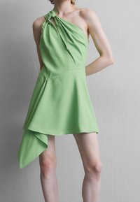 Grön asymmetrisk klänning gjord av mjukt tyg, med en knuten axeldetalj och utsvängd fåll, vilket skapar en svepande silhuett.