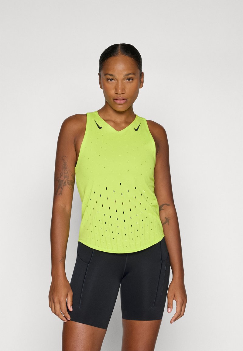 Canotta atletica verde lime con scollo a V, caratterizzata da piccole perforazioni per la ventilazione. Abbinata a pantaloni corti neri aderenti con tasche laterali.