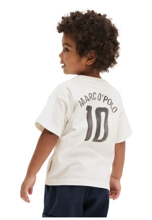 Tout-petit aux cheveux bouclés portant un t-shirt blanc Marc O’Polo avec le numéro 10 au dos et un short sombre, regardant sur le côté.