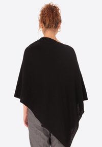 Poncho nero in maglia con forma triangolare, texture morbida e design drappeggiato. Indossato sopra pantaloni grigi, mette in mostra la sua silhouette fluida.