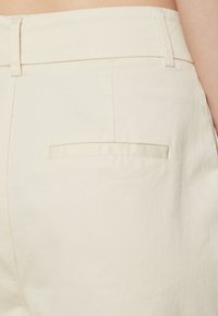 Pants svijetle boje krem s pamukom, s glatkom teksturom, ravnim stražnjim džepom i ušivenim detaljima duž pojasa i šavova.