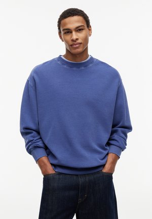 Ung mand iført en blå tekstureret langærmet trøje og mørke jeans med hænderne i lommerne, stående mod en ensfarvet hvid baggrund.