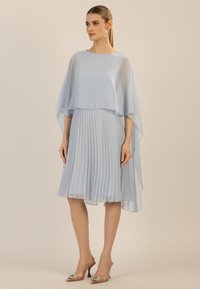 Robe plissée bleu clair avec une superposition de cape transparente, longueur genoux, associée à des chaussures à talons décoratives. Tissu lisse et texture douce.
