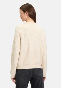 Cremefarbener Pullover mit einem strukturierten, erhabenen abstrakten Muster. Mit Rundhalsausschnitt und gerippten Bündchen. Kombiniert mit dunklen Jeans, von hinten betrachtet.