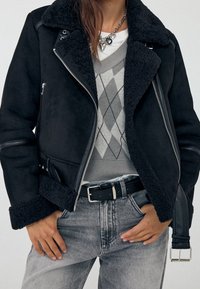 Vrouw in een zwarte schapenvacht jack met riem, grijze argyle trui, lichtgewassen jeans en een hartvormige hangersnoer, met handen in haar zakken.
