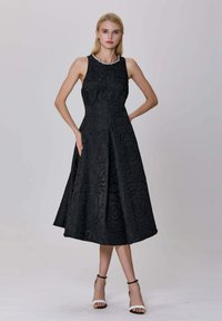 Femme blonde en robe noire sans manches avec des motifs floraux texturés et un collier de perles, portant des sandales à talons hauts blanches, se tenant les mains sur les hanches.